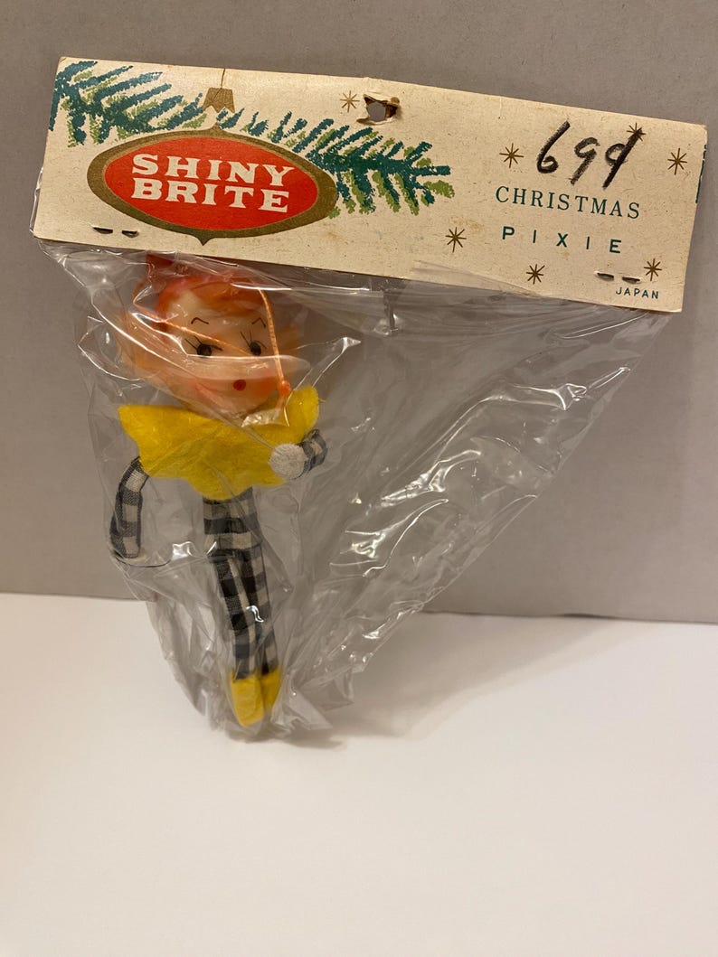 May include: A vintage Christmas pixie ornament in a plastic bag. The ornament has a yellow collar, black and white checkered pants, and yellow shoes. The packaging reads "Shiny Brite" and "Christmas Pixie."
