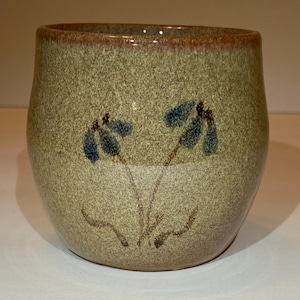 May include: A ceramic mug with a speckled beige and tan finish, featuring a hand-painted design of two blue flowers with brown stems. The rim of the mug has a distinct reddish-brown band, and the overall shape is rounded with a wide opening.