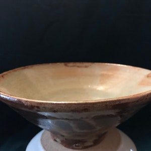 May include: A wide, shallow ceramic bowl with a light brown glaze. The bowl has a slightly flared rim and a rounded bottom. The glaze is uneven, with some areas of darker brown and white.