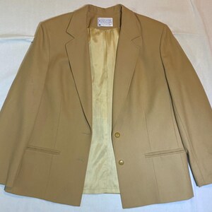 May include: A tan blazer with a two-button closure, two front pockets, and a notched lapel. The interior lining is a light yellow color. The blazer is made of a smooth fabric.