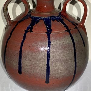 May include: A handcrafted ceramic vase with a round body and two loop handles. The vase is primarily red with gray and blue accents. Dark blue lines drip down the sides, adding a decorative touch. The vase has a small opening at the top.