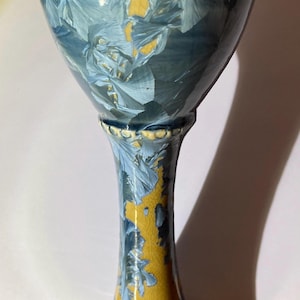 May include: A tall, handcrafted ceramic goblet with a flared top and a slender stem. The goblet features a striking blue and yellow crystalline glaze, with a light yellow rim. The design is reminiscent of ice crystals.