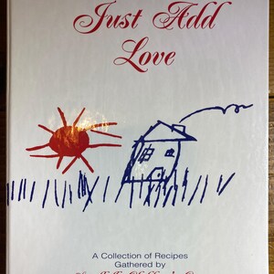 May include: A cookbook cover with the title "Just Add Love" in red script. The cover features a child's drawing of a red sun, a blue house with a smoking chimney, and blue grass. The text "A Collection of Recipes Gathered by Sandhills Children's Center" is also present.