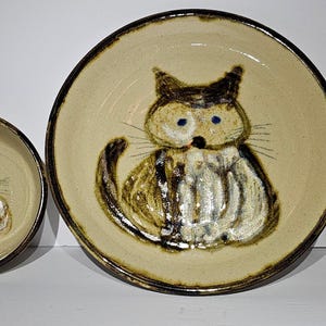 May include: A ceramic dish set featuring a cat design. The set includes a bowl, a plate, and a mug. Each piece has a beige background with a brown and white cat illustration. The cat has blue eyes and a whimsical style.