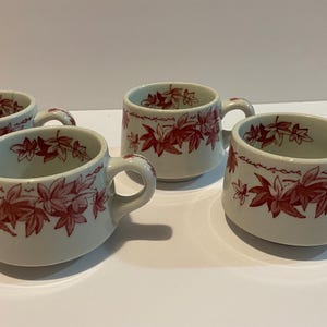 May include: Four white ceramic cups with handles, each adorned with a red leaf pattern. The cups have a slightly angled design and are arranged on a white surface.