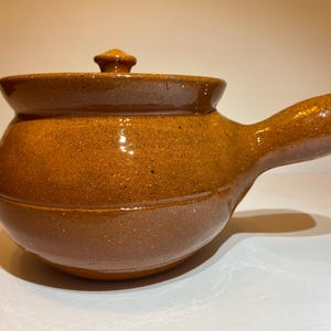 May include: A brown ceramic soup pot with a lid and a handle. The pot has a rounded body and a lid with a small knob. The handle is curved and extends from the side of the pot. The pot is glazed and has a glossy finish.