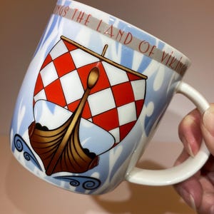 May include: White ceramic mug with a handle, featuring a Viking ship design with a red and white checkered sail. The mug has a blue and white background with the text "Kings the Land of Vikings" in red.