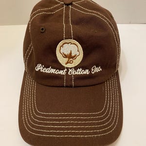 Puede incluir: Gorra de b&eacute;isbol marr&oacute;n con costuras blancas y visera curva. La gorra presenta el texto "Piedmont Cotton Inc." y un emblema circular con un dise&ntilde;o de planta de algod&oacute;n.