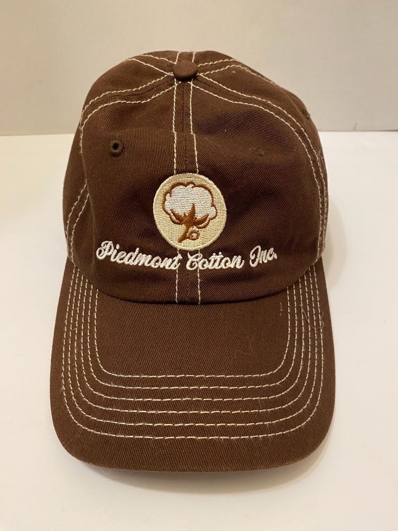 Puede incluir: Gorra de b&eacute;isbol marr&oacute;n con costuras blancas y visera curva. La gorra presenta el texto "Piedmont Cotton Inc." y un emblema circular con un dise&ntilde;o de planta de algod&oacute;n.