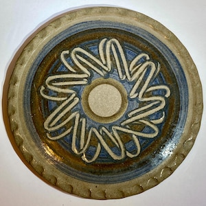 May include: A round ceramic plate featuring a central beige circle surrounded by a raised, white, looped floral pattern. The surface displays concentric rings of blue and brown glaze, with a textured, scalloped edge in a light tan finish.