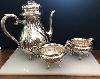 May include: A silver tea set with a teapot, creamer, and sugar bowl. The set has a classic design with ornate details and a polished finish.