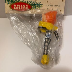 May include: A vintage Christmas pixie ornament in a plastic bag. The pixie has an orange hat, yellow jacket, and black and white plaid pants. The packaging reads "SHINY BRITE" and "CHRISTMAS PIXIE."