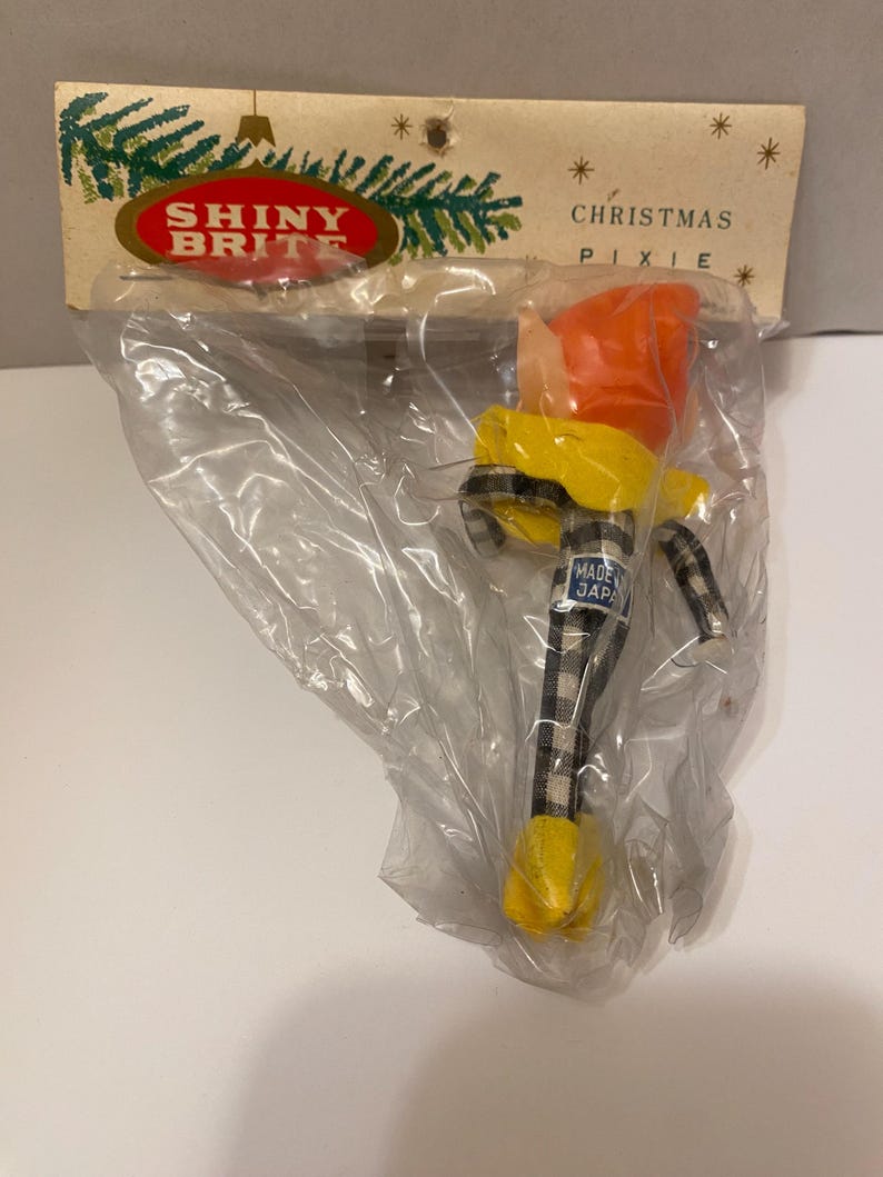 May include: A vintage Christmas pixie ornament in a plastic bag. The pixie has an orange hat, yellow jacket, and black and white plaid pants. The packaging reads "SHINY BRITE" and "CHRISTMAS PIXIE."