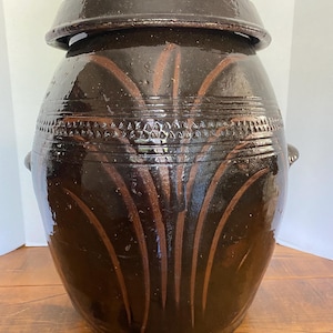 May include: A large, dark brown ceramic jar with a lid. The jar features decorative vertical and curved brown lines, and a band of textured detailing around the middle. Two small handles are on the sides.