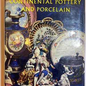 May include: A hardcover book titled The Concise Encyclopedia of Continental Pottery and Porcelain. The cover features a dark blue background with a collage of colorful ceramic items, including a blue vase, a decorative plate, and figurines.