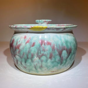 May include: A round ceramic lidded jar featuring a marbled glaze of mint green, white, and deep magenta. The lid has a matching marbled pattern and a central knob. A prominent gold-colored repair mark is visible on the rim of the jar.