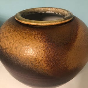May include: A large, round, brown ceramic vase with a textured surface. The vase has a light brown rim and a dark brown base.