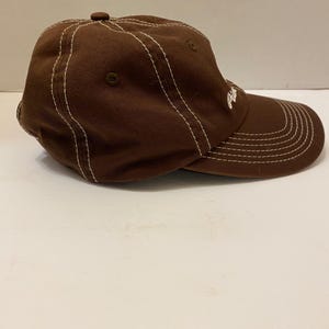 Puede incluir: Gorra de b&eacute;isbol marr&oacute;n con costuras blancas. La gorra tiene una visera curva y un bot&oacute;n en la parte superior. Un peque&ntilde;o logotipo es visible en el lateral.