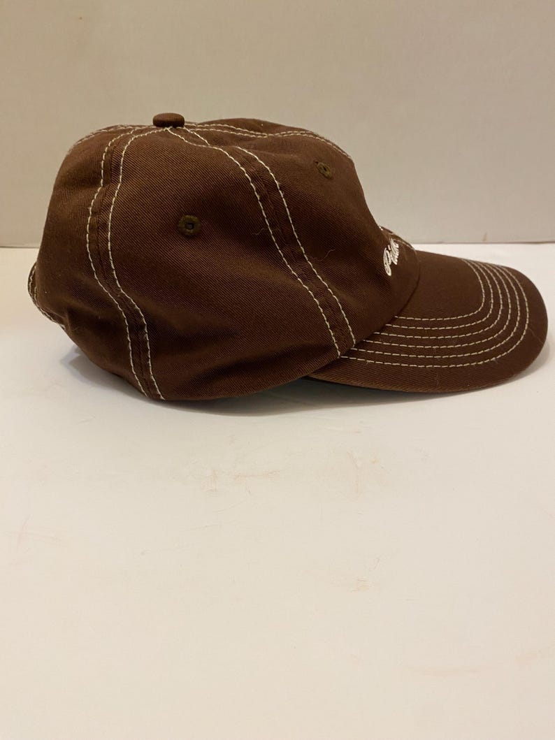 Puede incluir: Gorra de b&eacute;isbol marr&oacute;n con costuras blancas. La gorra tiene una visera curva y un bot&oacute;n en la parte superior. Un peque&ntilde;o logotipo es visible en el lateral.