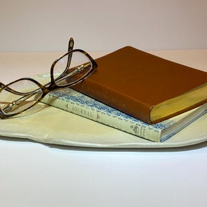 May include: A light-colored, oval-shaped ceramic tray holds a pair of tortoiseshell eyeglasses, a brown leather-bound book, and a blue and white patterned journal. The book has gold-colored edges.