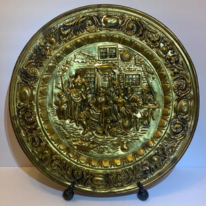 May include: A round brass decorative plate featuring a high-relief embossed scene of a group of people gathered outside a building. The border is decorated with intricate scrollwork and floral patterns, with a polished gold-toned metallic finish.