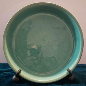 May include: A large, round, turquoise ceramic plate with a smooth, glossy finish. The plate is sitting on two black metal stands.