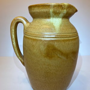May include: A ceramic pitcher with a handle and a spout. The pitcher is a warm, earthy tone of brown with a glossy finish. Two horizontal lines encircle the body of the pitcher. The pitcher is approximately 9 inches tall.