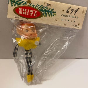 May include: A vintage Christmas pixie ornament in a clear plastic bag. The pixie has orange hair, glasses, and a yellow collar. It wears black and white checkered pants and yellow shoes. The packaging reads "SHINY BRITE" and "CHRISTMAS PIXIE".