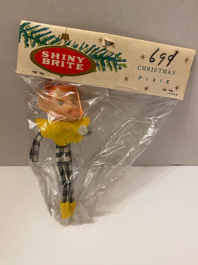 May include: A vintage Christmas pixie ornament in a clear plastic bag. The pixie has orange hair, glasses, and a yellow collar. It wears black and white checkered pants and yellow shoes. The packaging reads "SHINY BRITE" and "CHRISTMAS PIXIE".