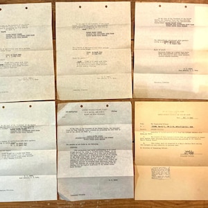 May include: A collection of vintage documents, including letters and official forms, are arranged on a wooden surface. The papers are aged with visible creases and text, suggesting historical significance. The documents are in shades of beige and cream.