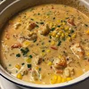 May include: A creamy, golden-colored seafood chowder in a white and blue ceramic pot. The chowder contains chunks of seafood, corn kernels, and green herbs. The pot sits on a white surface.