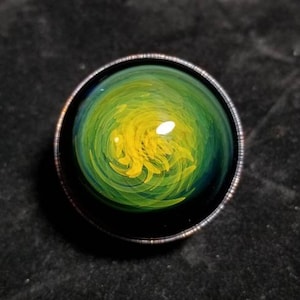May include: A glass marble with a swirling green and yellow design. The marble is encased in a black metal ring.