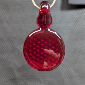 May include: A red glass pendant with a round, dotted design and a teardrop-shaped top. The pendant is suspended from a thin, beige cord. The background is a dark, textured surface.