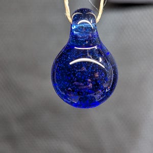 May include: A teardrop-shaped, deep blue glass pendant with a speckled interior. The pendant is suspended by a natural fiber cord. The glass has a glossy finish and appears to have small air bubbles within.