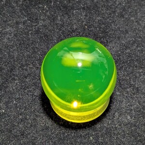 May include: A translucent green and yellow glass object, possibly a decorative knob or stopper. The top is a rounded dome shape, with a bright green hue. The base is a lighter yellow color. The object is set against a dark, textured background.