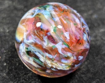May include: A colorful glass marble with swirling patterns of red, orange, blue, green, and white. The marble is round and appears to be handmade, with a glossy finish. The marble is set against a dark, textured background.