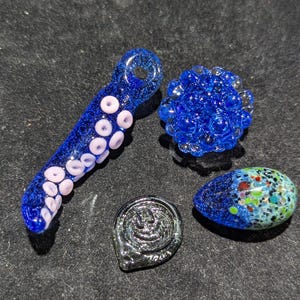 May include: An assortment of glass art pieces in shades of blue, including a pipe with white accents, a round cluster, a teardrop shape with colorful speckles, and a small, silver-toned piece.