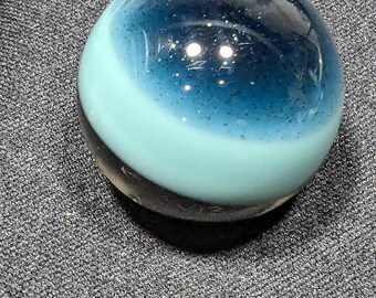 May include: A clear glass marble with a layered design. The top half is a deep blue with glitter, transitioning to a light blue band, and ending with a dark base. The marble is resting on a gray textured surface.