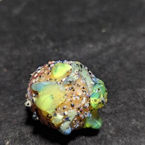 May include: A colorful glass bead with a textured surface. The bead features a mix of colors, including yellow, green, blue, and brown, with small, speckled details. The overall shape is rounded and organic.