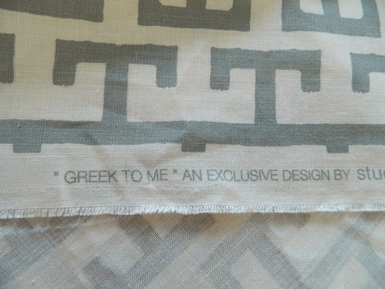 May include: Close-up of fabric with a repeating gray Greek key pattern on a cream background. The text "GREEK TO ME" is printed along the bottom edge. An exclusive design.