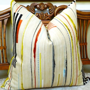 May include: A decorative throw pillow with a cream-colored background and vertical stripes in black, orange, red, yellow, and gray. The pillow is placed on a wooden chair with an ornate back.
