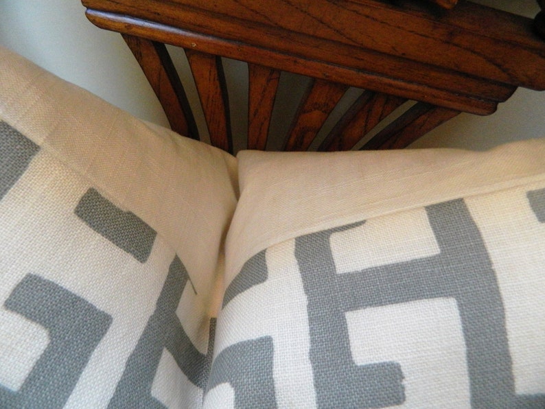 May include: Two decorative throw pillows with a white background and a gray geometric pattern. The pillows are rectangular and have a light-colored border. A wooden decorative element is visible in the background.