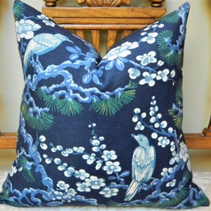 May include: A square decorative pillow with a navy blue background. The pillow features a detailed print of birds, branches, and flowers in shades of blue, white, and green. The pillow is placed on a wooden chair.
