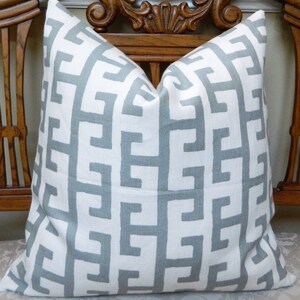 May include: Square decorative pillow with a white background and a repeating geometric pattern in a light gray color. The pillow is made of a textured fabric and is placed on a wooden chair.