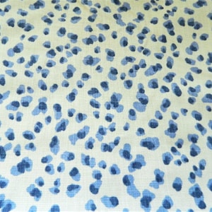 May include: Close-up of a fabric with a repeating pattern of blue, irregular shapes against a cream-colored background. The shapes vary in size and density, creating a speckled appearance. The fabric has a slightly textured surface.