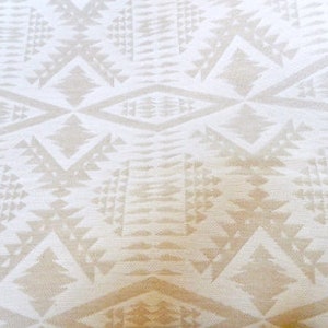 May include: Cream-coloured fabric with a repeating geometric pattern. The design features diamond and triangle shapes in a lighter shade of cream, creating a textured appearance. The fabric has a soft, slightly blurred look.