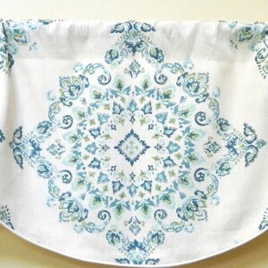 Peut inclure: Une cantonnière blanche avec un motif floral bleu et vert répété. La cantonnière a un bord inférieur festonné et est froncée en haut pour être suspendue. Le motif est symétrique, avec un médaillon central et des accents d'angle.