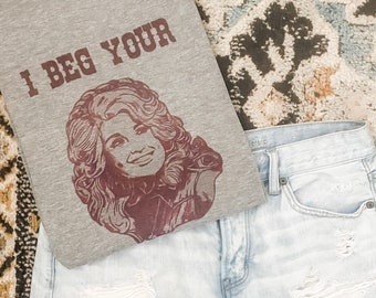 May include: A gray t-shirt with a brown print of a woman with long blonde hair. The text "I beg your" is printed on the shirt.