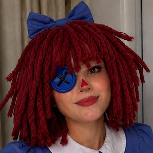 Pode incluir: Um traje inspirado em uma boneca com um vestido azul de gola e punhos brancos, adornado com remendos de tecido azul escuro. O visual inclui uma peruca de lã vermelha com um laço azul, um botão azul grande sobre um olho e um nariz triangular vermelho.