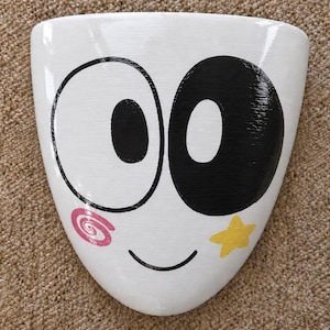 Puede incluir: Una maceta de pared de cerámica blanca con forma triangular que presenta una cara de dibujos animados con un ojo grande negro y un ojo grande blanco. La cara incluye una pequeña sonrisa negra curvada, una marca de espiral rosa y una estrella amarilla.